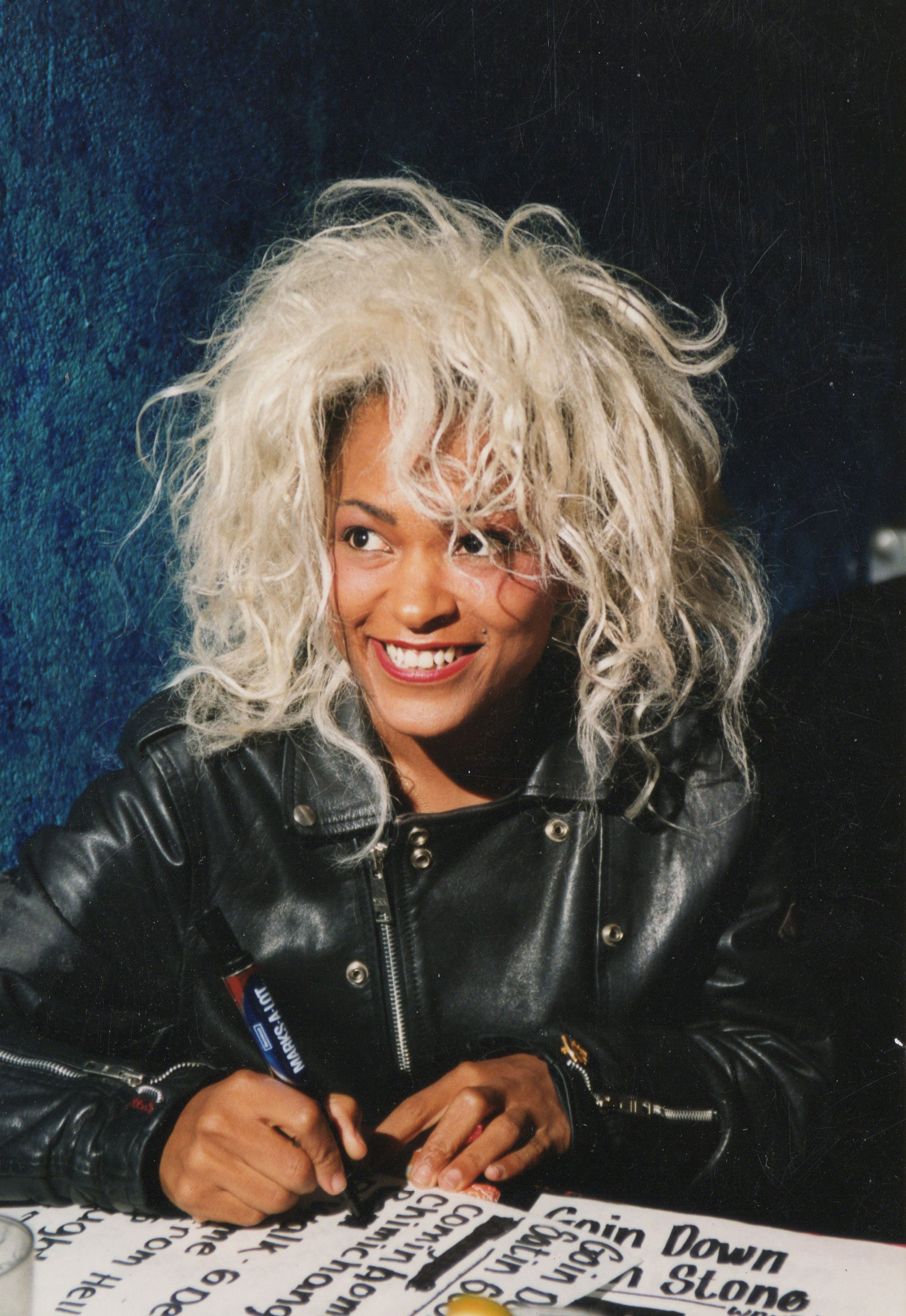 Person wearing black leather jacket smiles, their hair is platinum blonde. They are signing something on the table in front of them with a thick black marker.