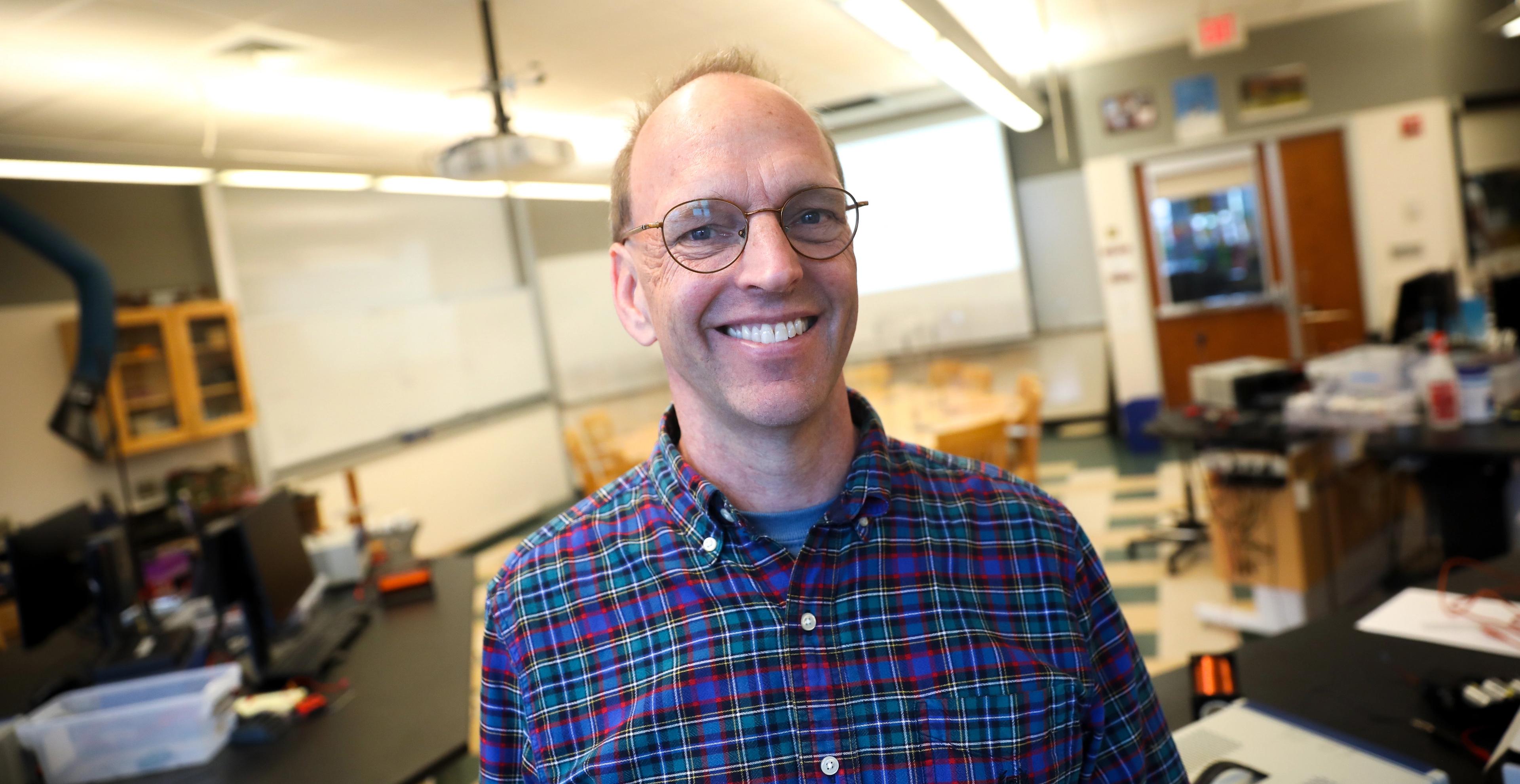 A teacher stands in his classroom and grins.