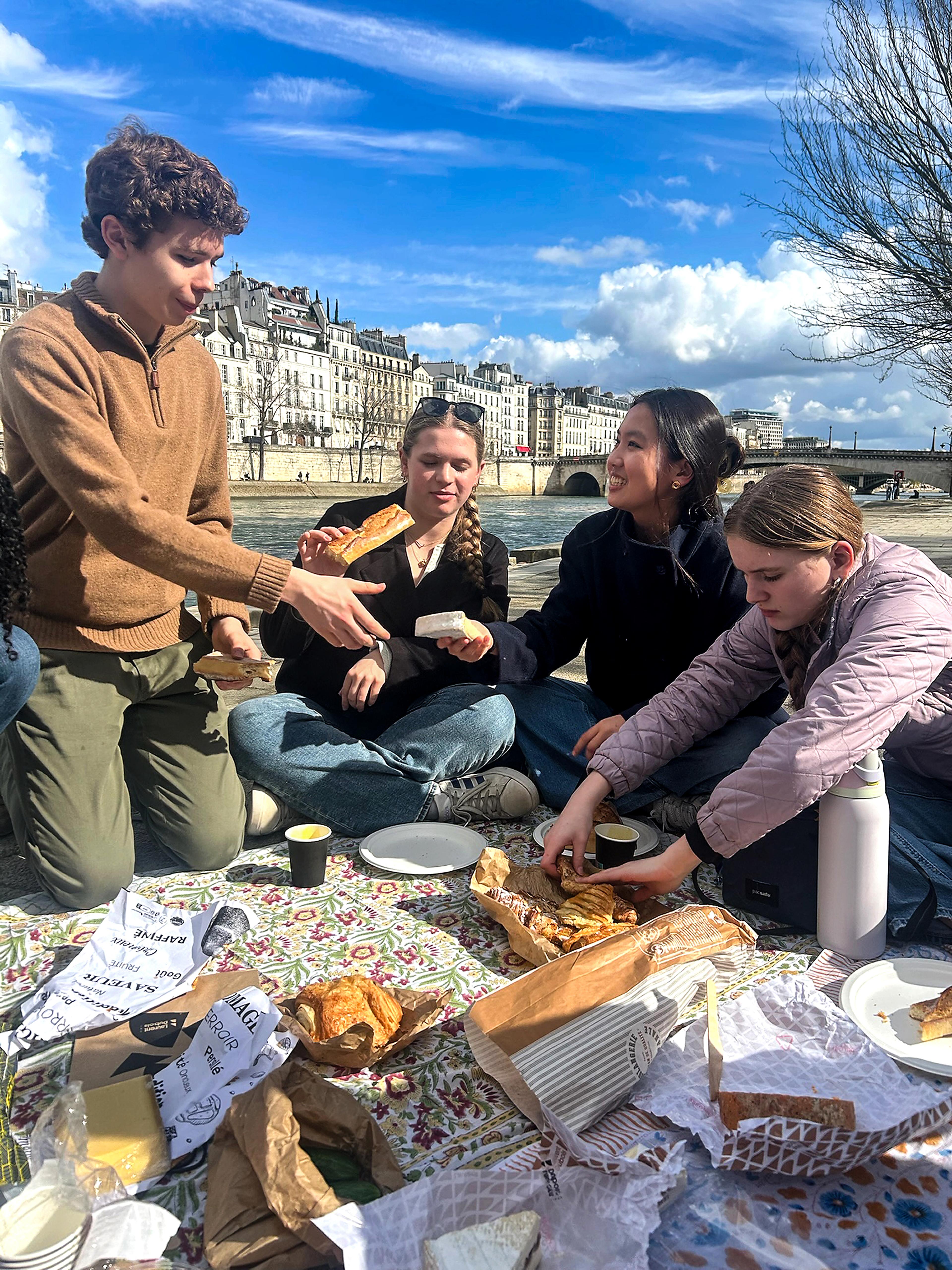 Teenagers having a picnic by a river with building behind 