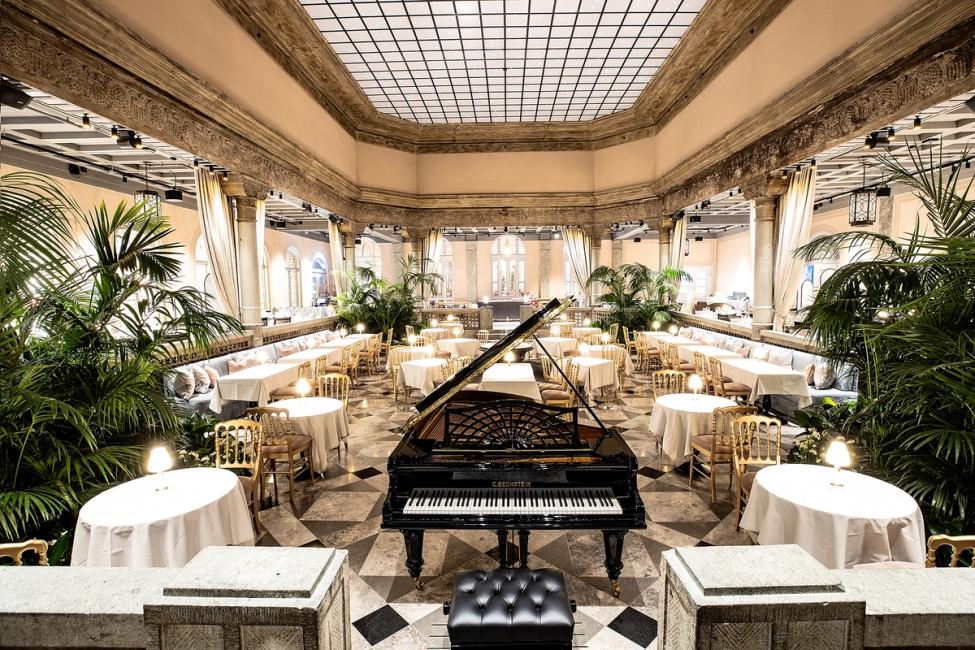 A grand ballroom with a black Bechstein piano, round white-clothed tables, gold chairs, and palm plants under a skylight — by Nonspace