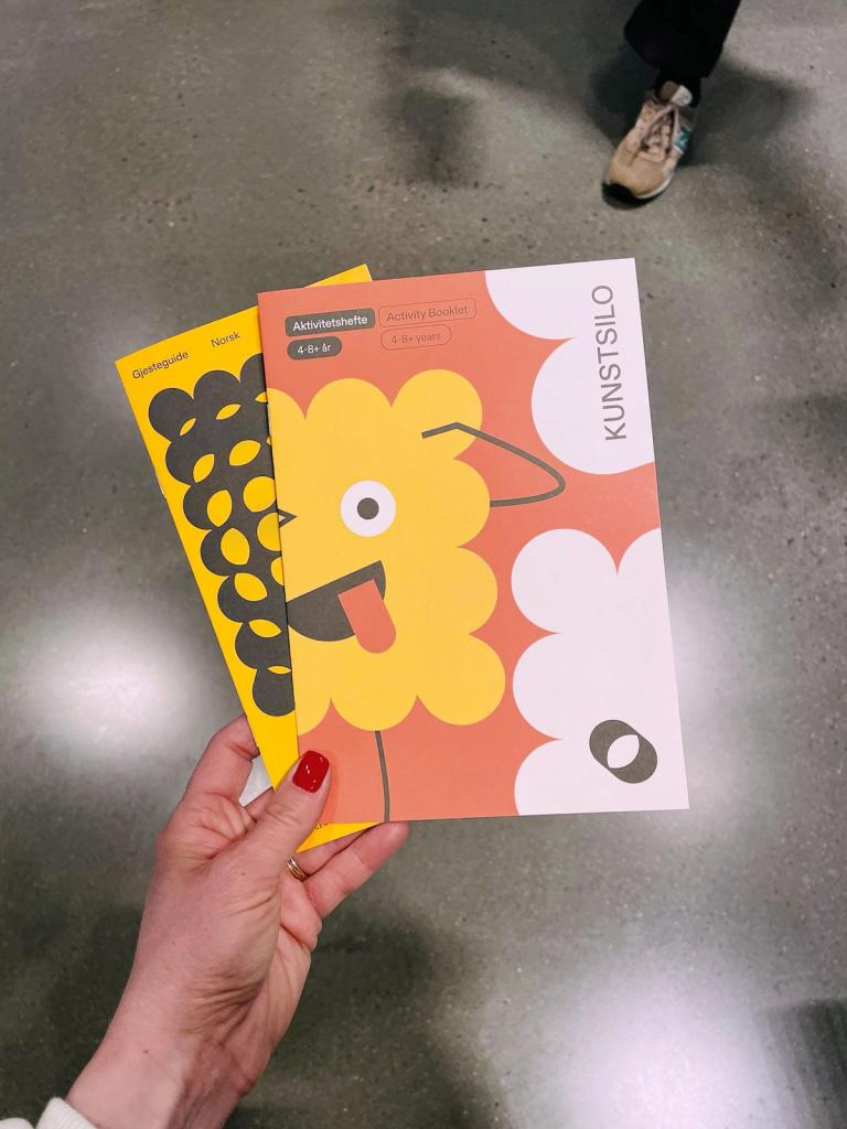 Hand holding two colourful children’s activity booklets from Kunstsilo, featuring playful illustrated characters, inside the museum’s kids area.