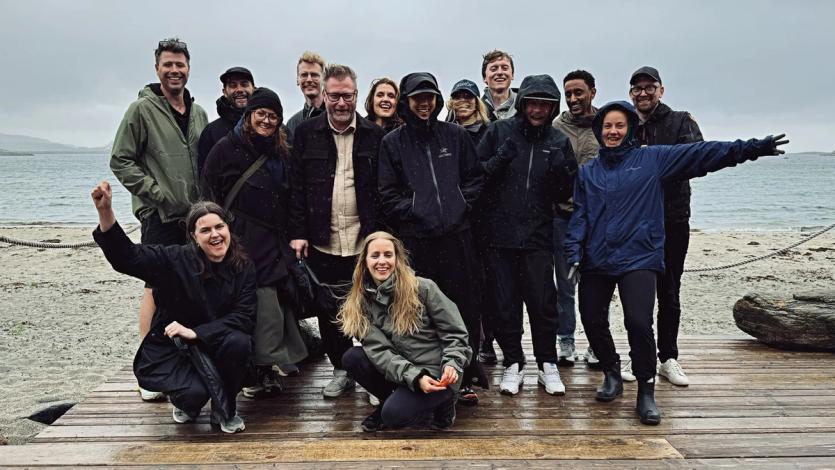 The Nonspace team gathered on a rainy beach at Stokkøya, smiling in rain gear while posing on a wooden boardwalk during the annual summer party.