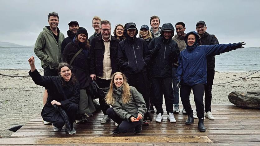The Nonspace team gathered on a rainy beach at Stokkøya, smiling in rain gear while posing on a wooden boardwalk during the annual summer party.