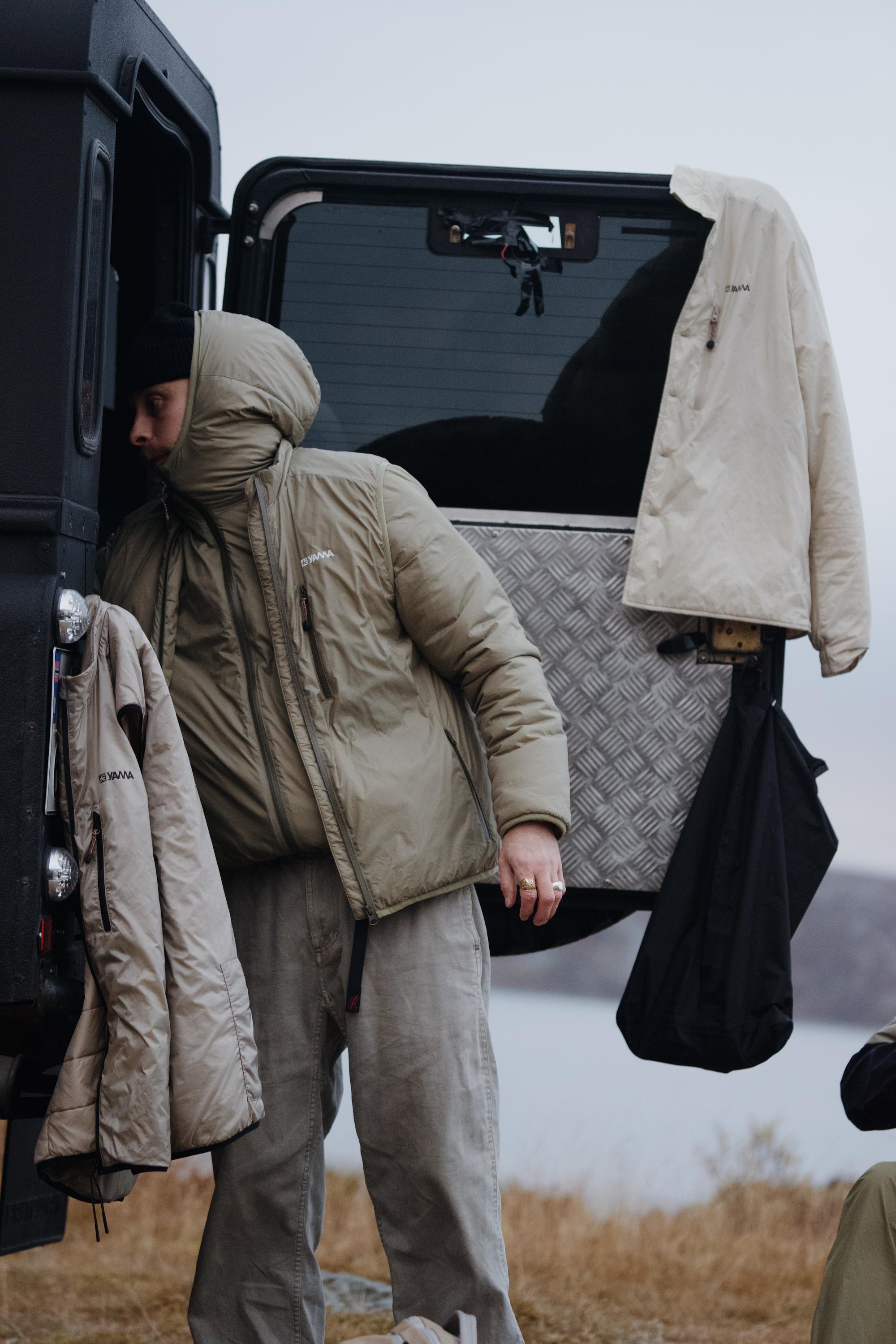 Person in a hooded Yama jacket leaning into the back of an off-road vehicle, with additional Yama outerwear hanging from the open door in a cold outdoor setting.