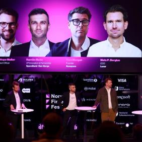 Four people on stage, audience in front, standing under large screen.