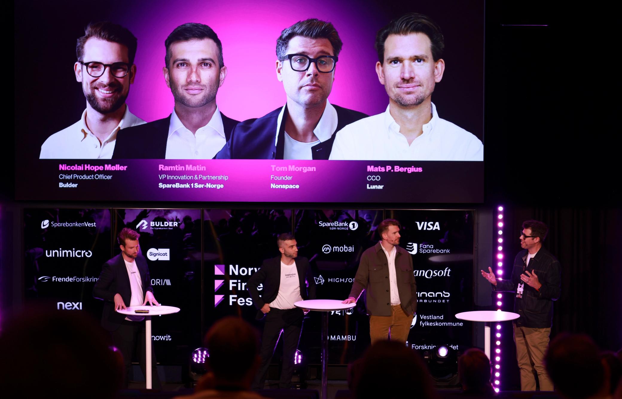 Four people on stage, audience in front, standing under large screen.