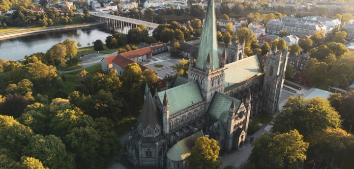 Aerial view of Nidaros Cathedral surrounded by trees and the city of Trondheim at sunset.