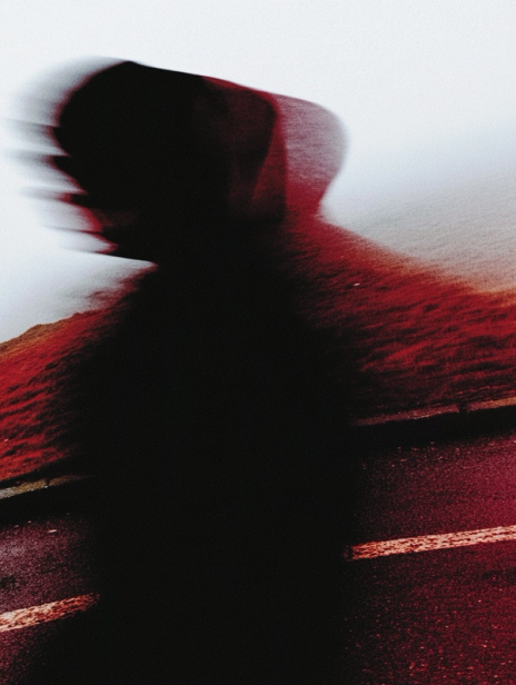 Blurred silhouette of a hooded person in motion on a road, with red tones blending into a misty landscape in the background.