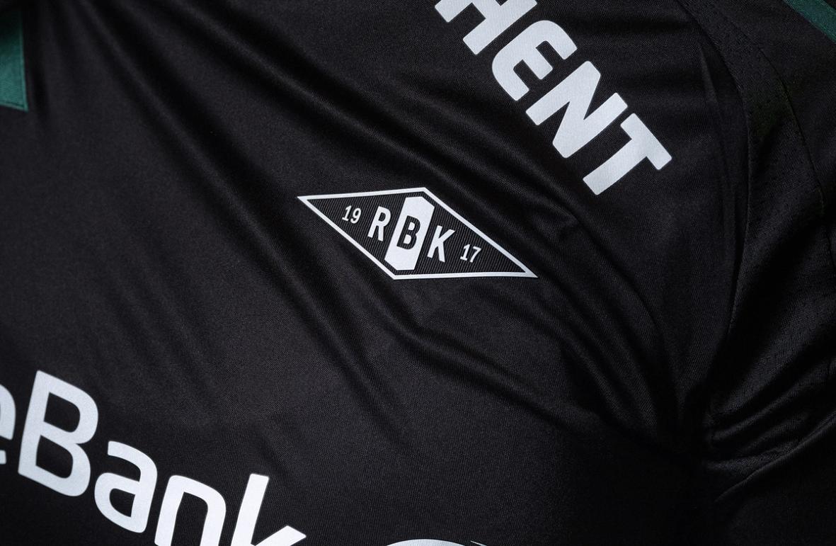 Close-up of a black Rosenborg football jersey showing the RBK crest printed on the chest, with white sponsor text visible on the fabric.
