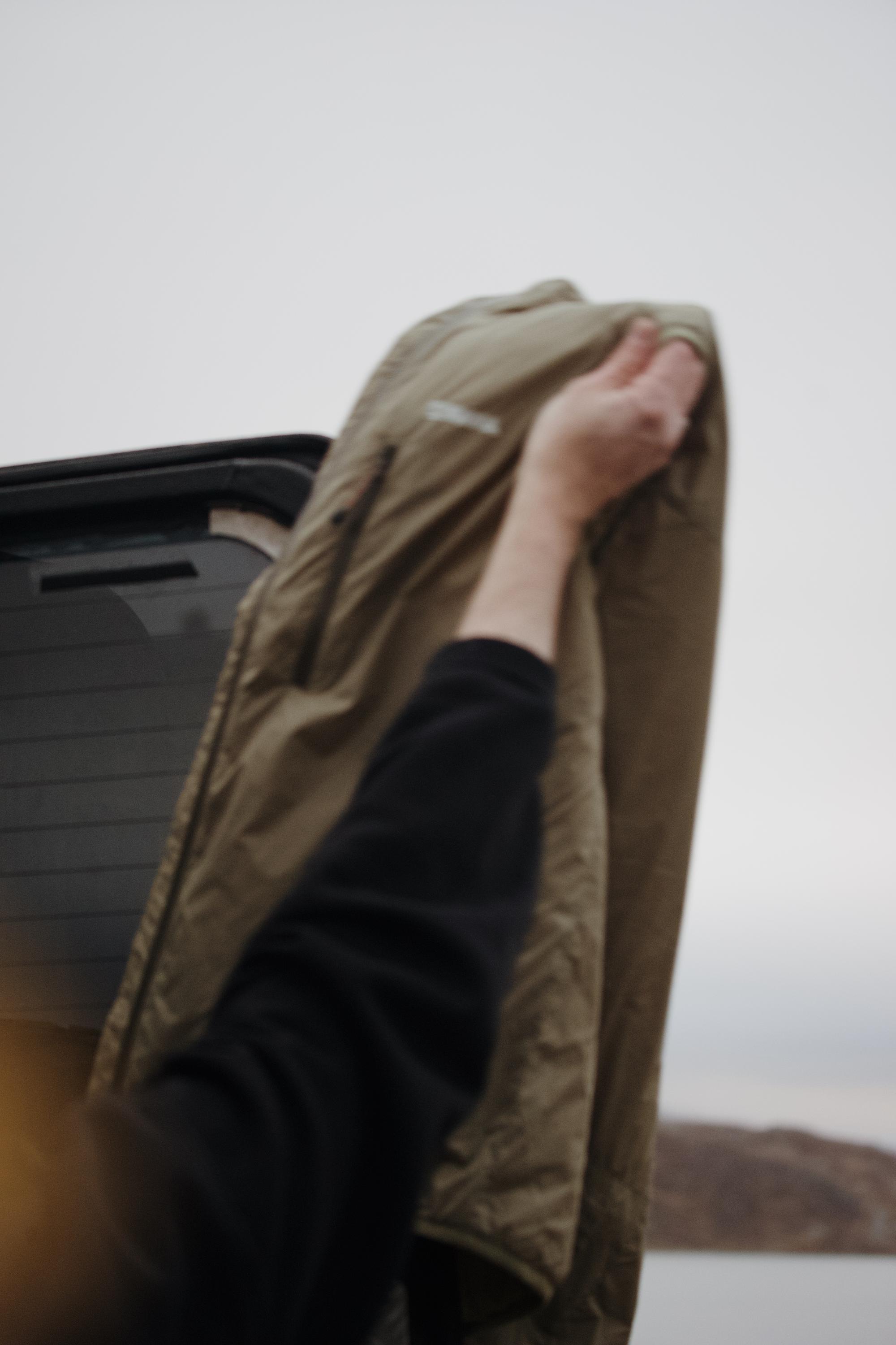Person lifting a beige Yama outdoor jacket beside a rugged off-road vehicle in a Nordic landscape, showcasing durable outdoor clothing for cold-weather adventures.