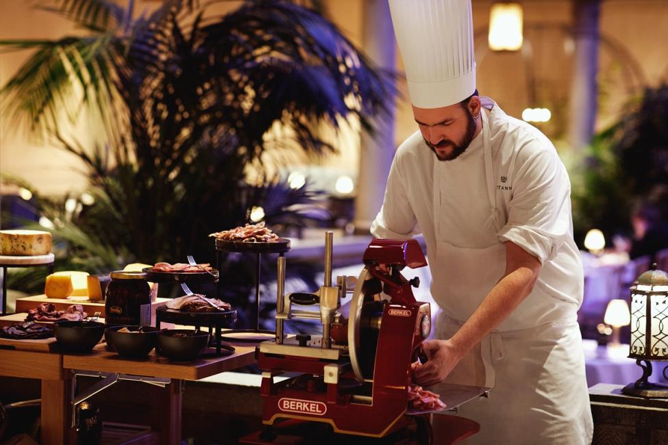 A chef in white uniform uses a red Berkel meat slicer at an elegant buffet station with cured meats and cheeses — by Nonspace