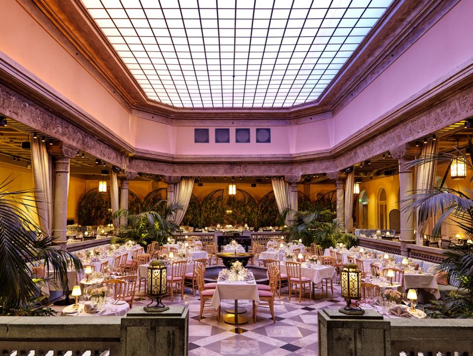 A grand hotel ballroom with elegant round tables, pink chairs, palm plants, and a large gridded skylight ceiling — by Nonspace