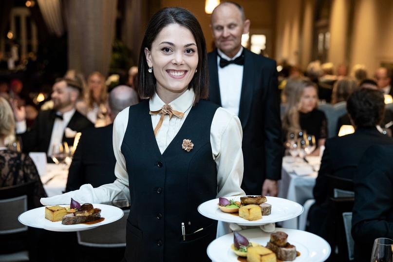 A smiling server in a black vest carries elegantly plated dishes at a formal gala dinner event — by Nonspace