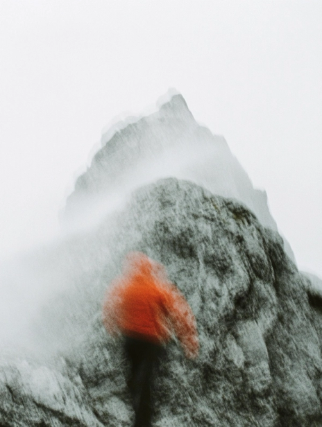 Blurred hiker in an orange jacket climbing a rugged grey mountain in windy, misty conditions, capturing outdoor adventure and nature movement.