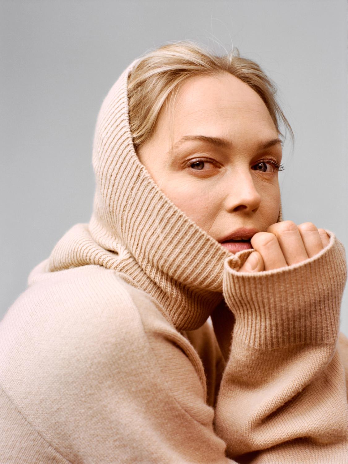 Close-up portrait of a woman wearing a soft beige knitted sweater with the hood pulled up, holding the collar near her face, showcasing cozy textured knitwear.