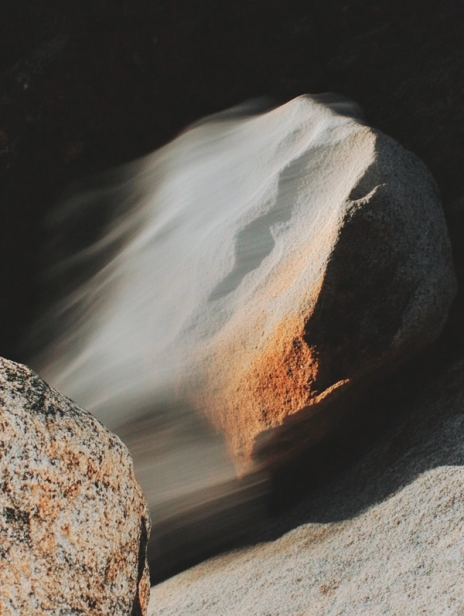 Wind blowing sand across sunlit rocks, creating soft, flowing motion over their textured surface.