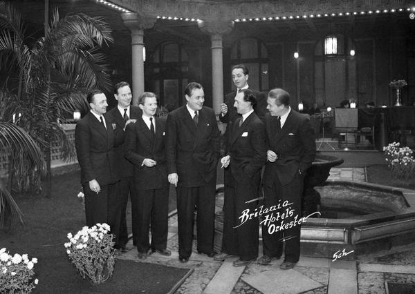 Seven men in suits gathered in an elegant hotel atrium with palm trees and a fountain — by Nonspace