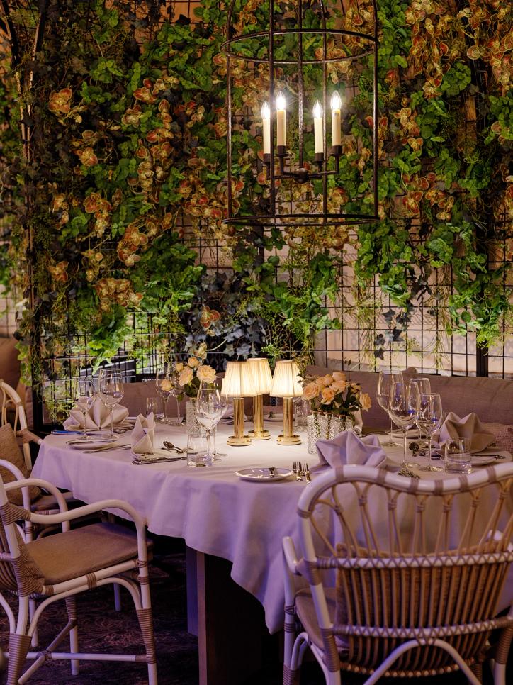 An elegant restaurant table setting surrounded by lush hanging greenery with a chandelier and soft table lamps — by Nonspace