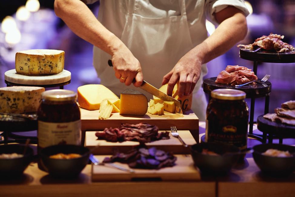 A chef slicing artisan cheese at an elegant charcuterie station with cured meats and preserves — by Nonspace