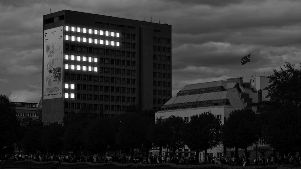 A tall building displays illuminated windows forming a pattern alongside a large festival poster at dusk — by Nonspace