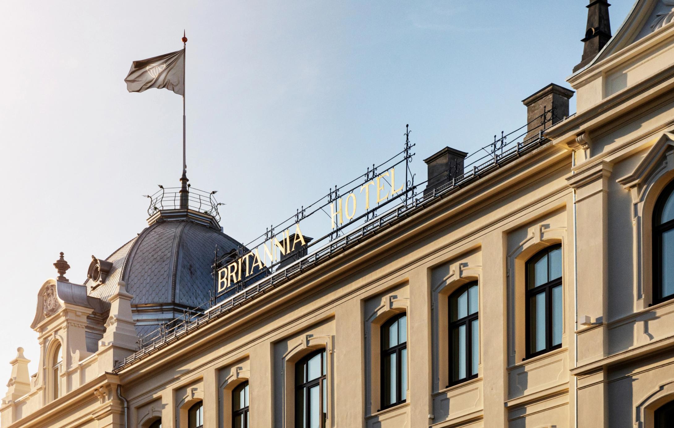 Britannia Hotels gule fasade med gullbokstaver på taket og flagg mot blå himmel — av Nonspace