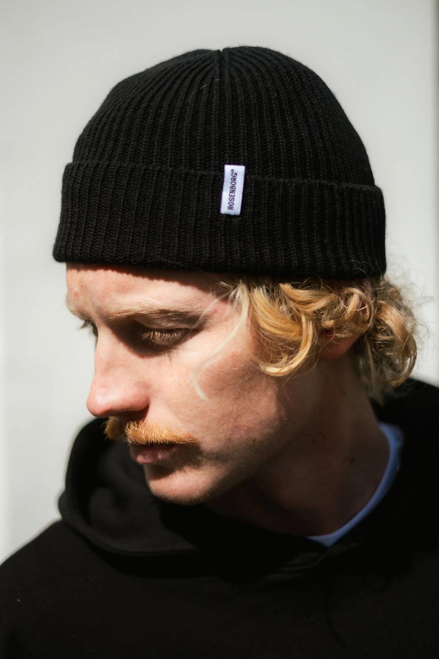En ung mann med blondt krøllete hår og bart ser ned, iført svart Rosenborg-lue og hettegenser — av Nonspace