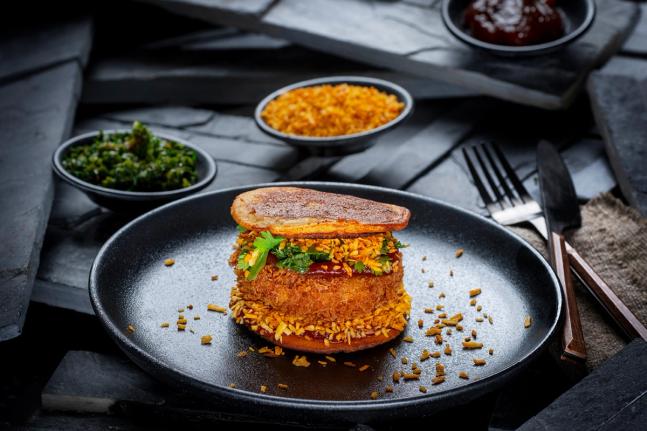 A crispy breaded sandwich with herbs on a black plate with sides of greens and rice — by Nonspace