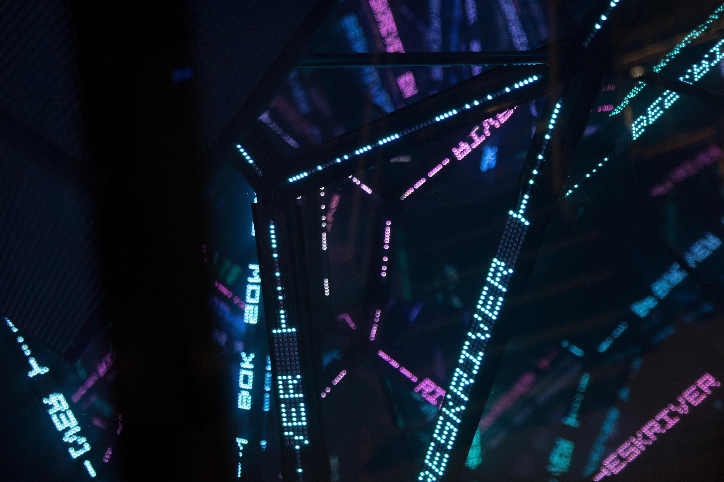 Lysende LED-tekst i cyan og magenta reflekteres i speilflater i et mørkt rom — av Nonspace