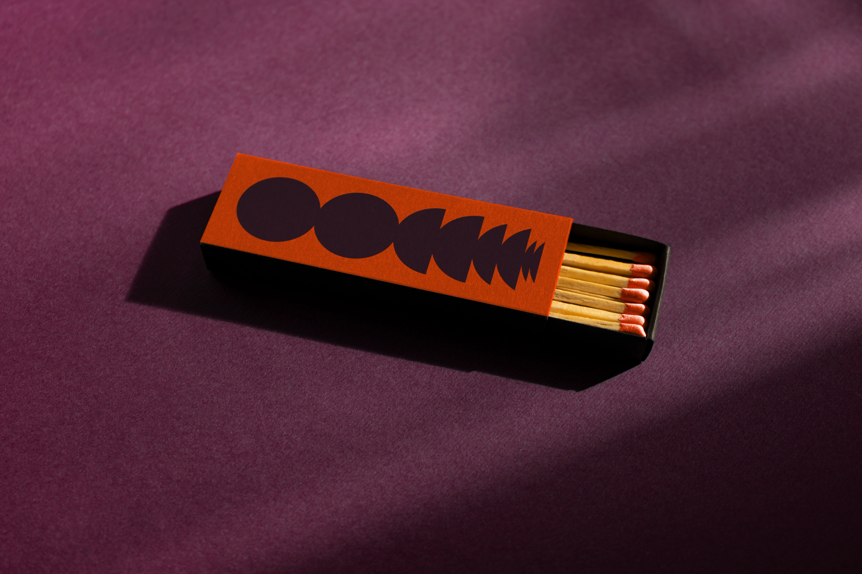 A matchbox with orange cover featuring abstract purple shapes, partially open to reveal wooden matches with red tips — by Nonspace