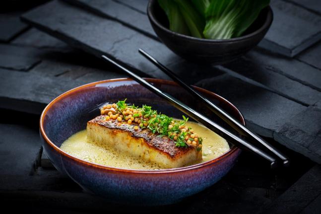 A seared fish fillet in golden broth topped with microgreens and crispy pearls in a ceramic bowl with chopsticks — by Nonspace