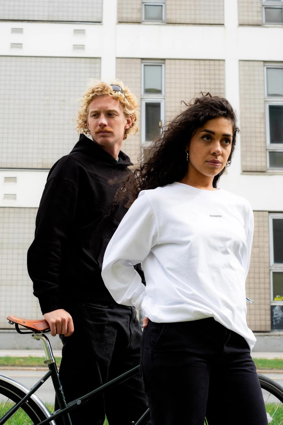 En mann og en kvinne i uformelle klær står ved en sykkel foran en bygård — av Nonspace