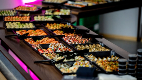 Et langbord med mange svarte fat fylt med variert sushi under rosa belysning — av Nonspace