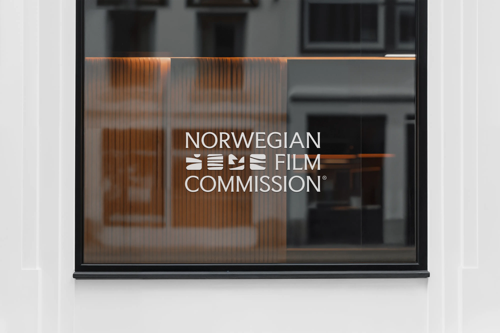 Vindu med Norwegian Film Commission-logo i hvit tekst på glass med trelameller synlige innenfor — av Nonspace