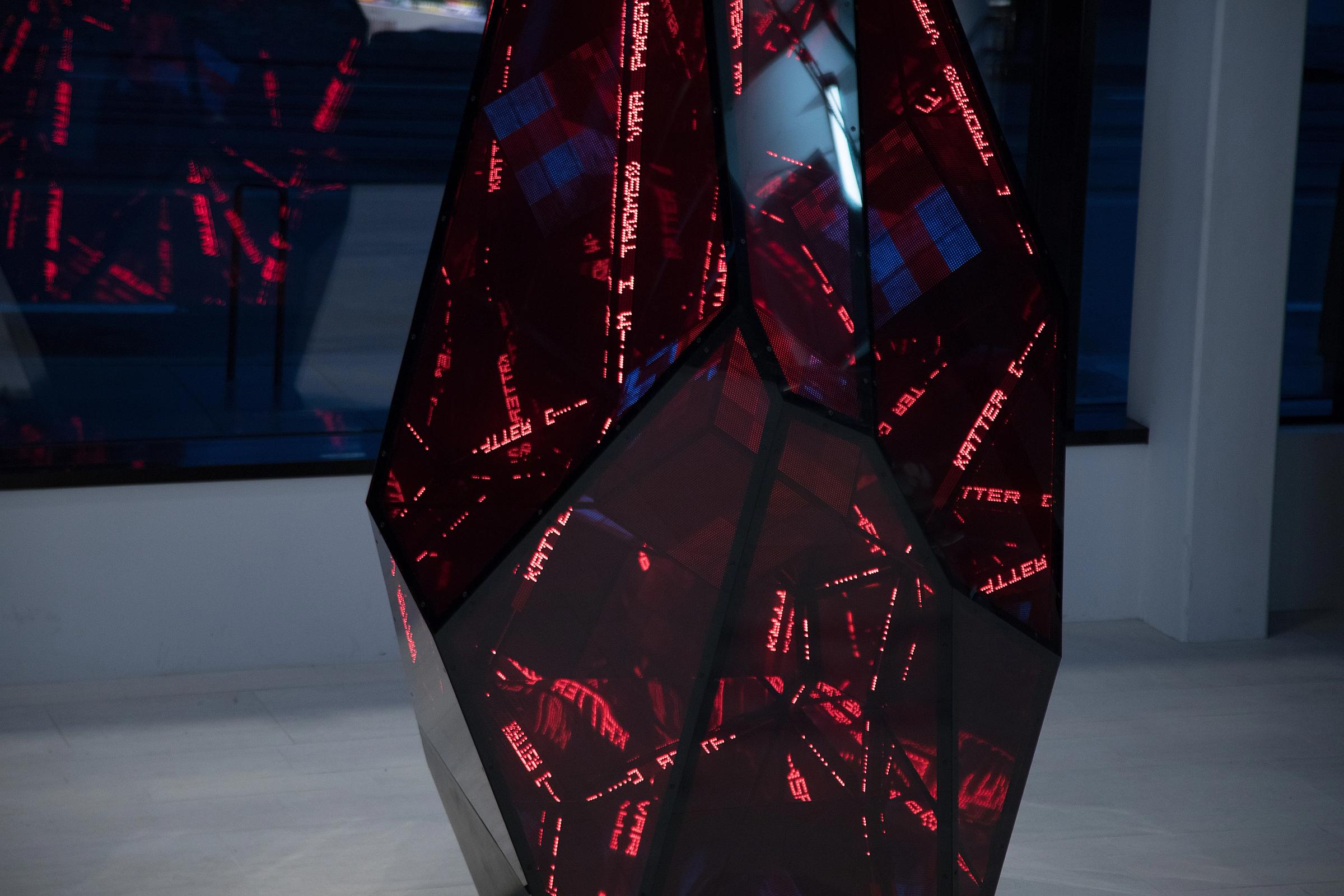 En flersidete skulptur med mørke, reflekterende flater og røde LED-tekstlinjer som lyser i et gallerirom — av Nonspace