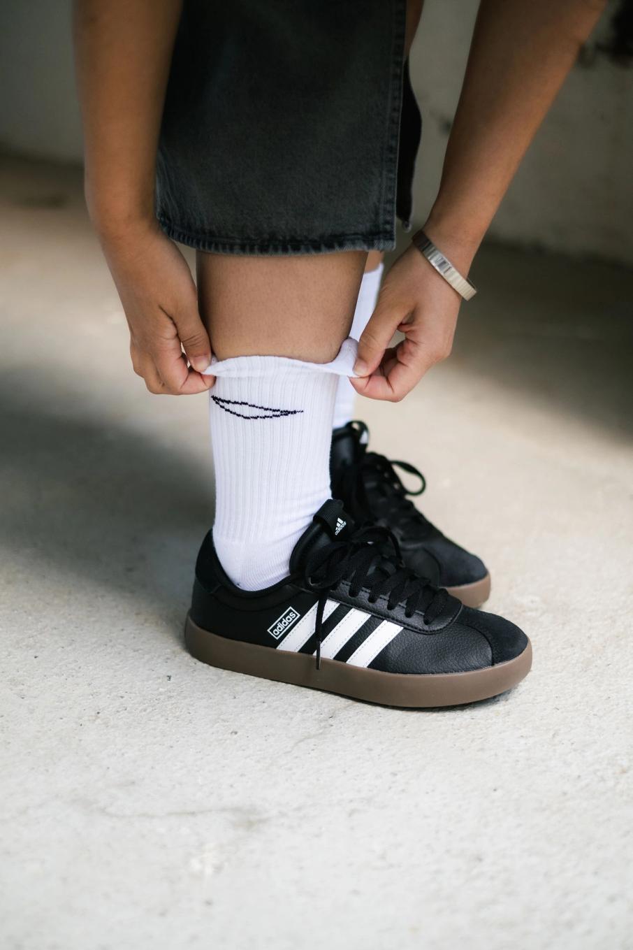 En person bøyer seg ned for å dra opp en hvit sokk over ankelen mens de har på svarte Adidas-sko — av Nonspace