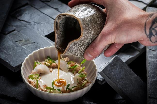 A tattooed hand pours amber broth from a ceramic vessel onto a plated dish with garnishes — by Nonspace