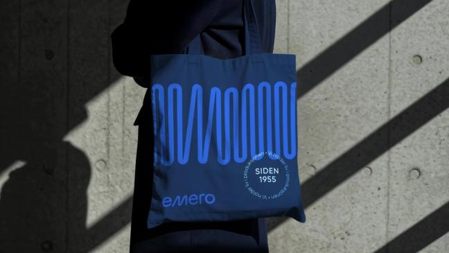 En person bærer en mørkeblå stoffpose med Emero-logo og bølgemønster i lyseblått mot en betongvegg — av Nonspace