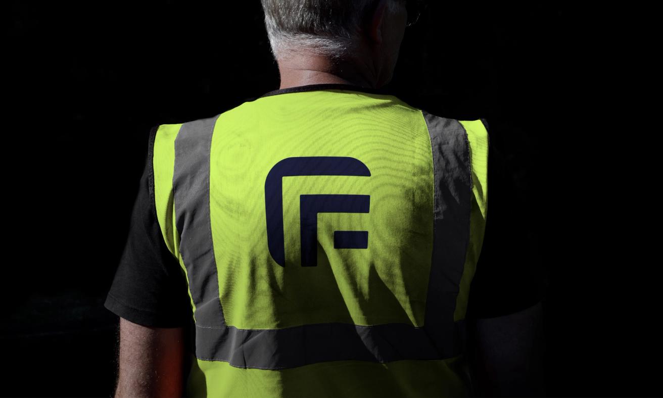 A worker's back wearing a yellow high-visibility safety vest with a navy F logo and reflective strips — by Nonspace