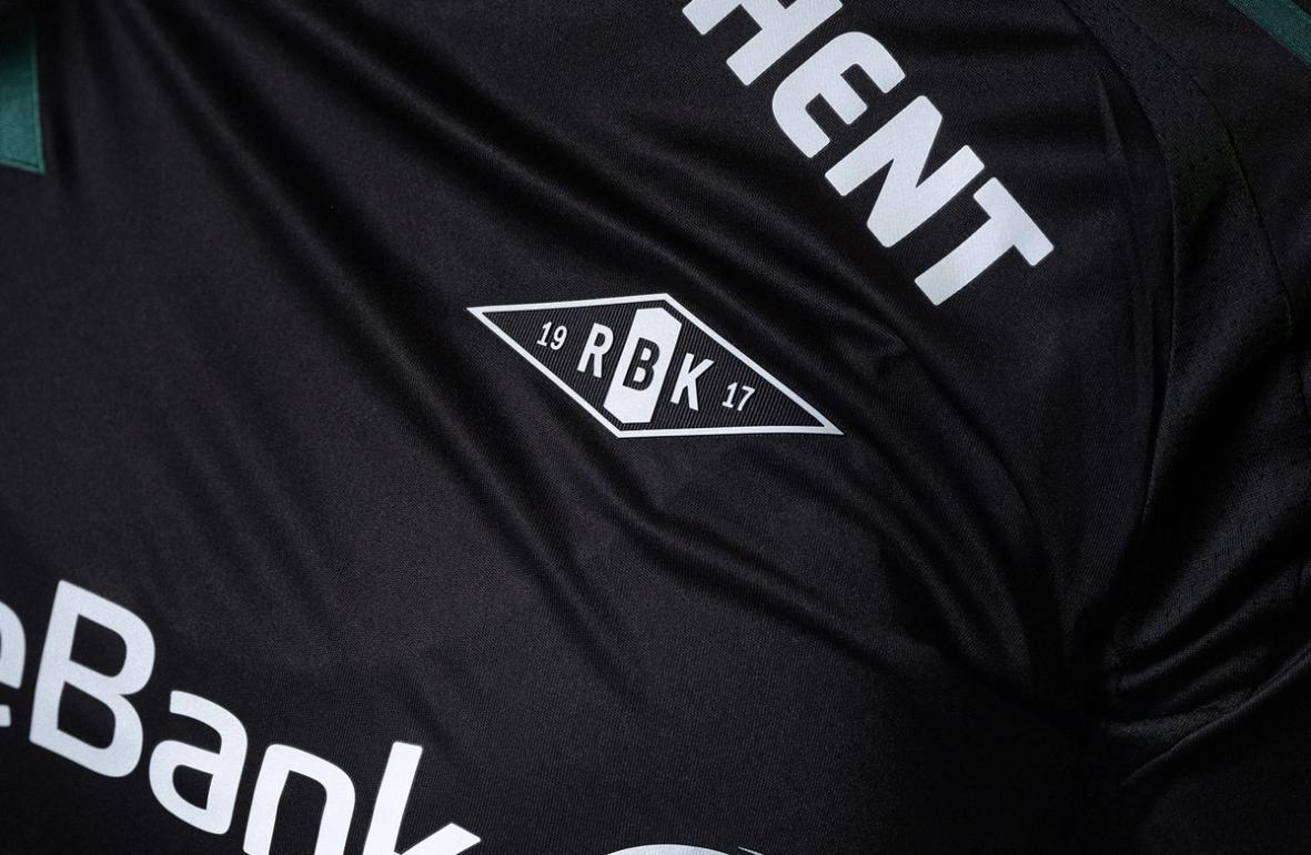 Close-up of a black Rosenborg football jersey showing the RBK crest printed on the chest, with white sponsor text visible on the fabric.