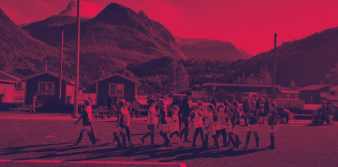 Children playing football on a field near small cabins with dramatic mountains in the background — by Nonspace