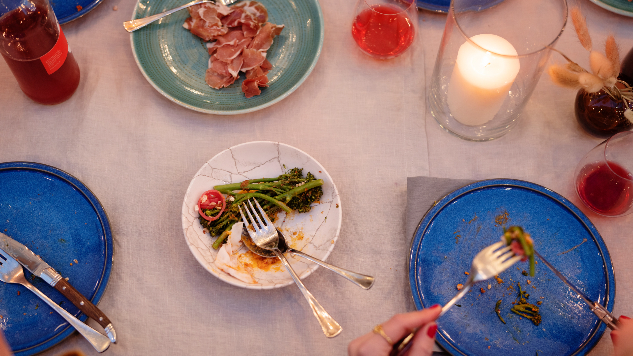 Et middagsbord med tallerkener med brokkolini og spekemat, vinglass, stearinlys og en hånd som spiser — av Nonspace