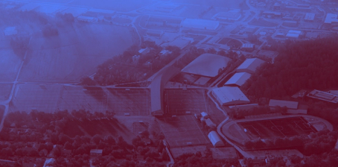 Flyfoto av et idrettsanlegg med fotballbaner, stadion og parkeringsplasser omgitt av skog og bebyggelse — av Nonspace