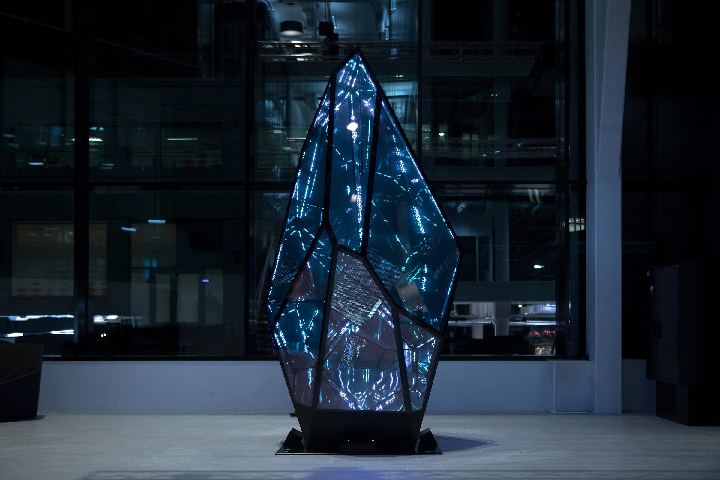 En krystallformet lysinstallasjon med blått LED-lys og geometriske glasspaneler i et mørkt gallerirom — av Nonspace