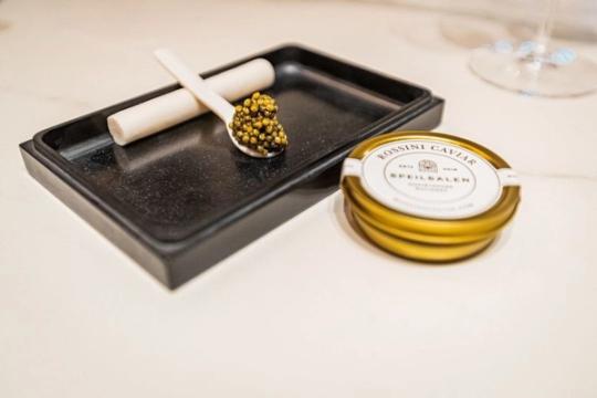 En skje med gyllen kaviar på svart skifer ved siden av en gullfarget Rossini Caviar-boks fra Speilsalen — av Nonspace