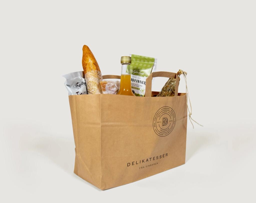 En brun papirpose med teksten Delikatesser fra Vinbaren fylt med baguett, juice, pølser og snacks — av Nonspace