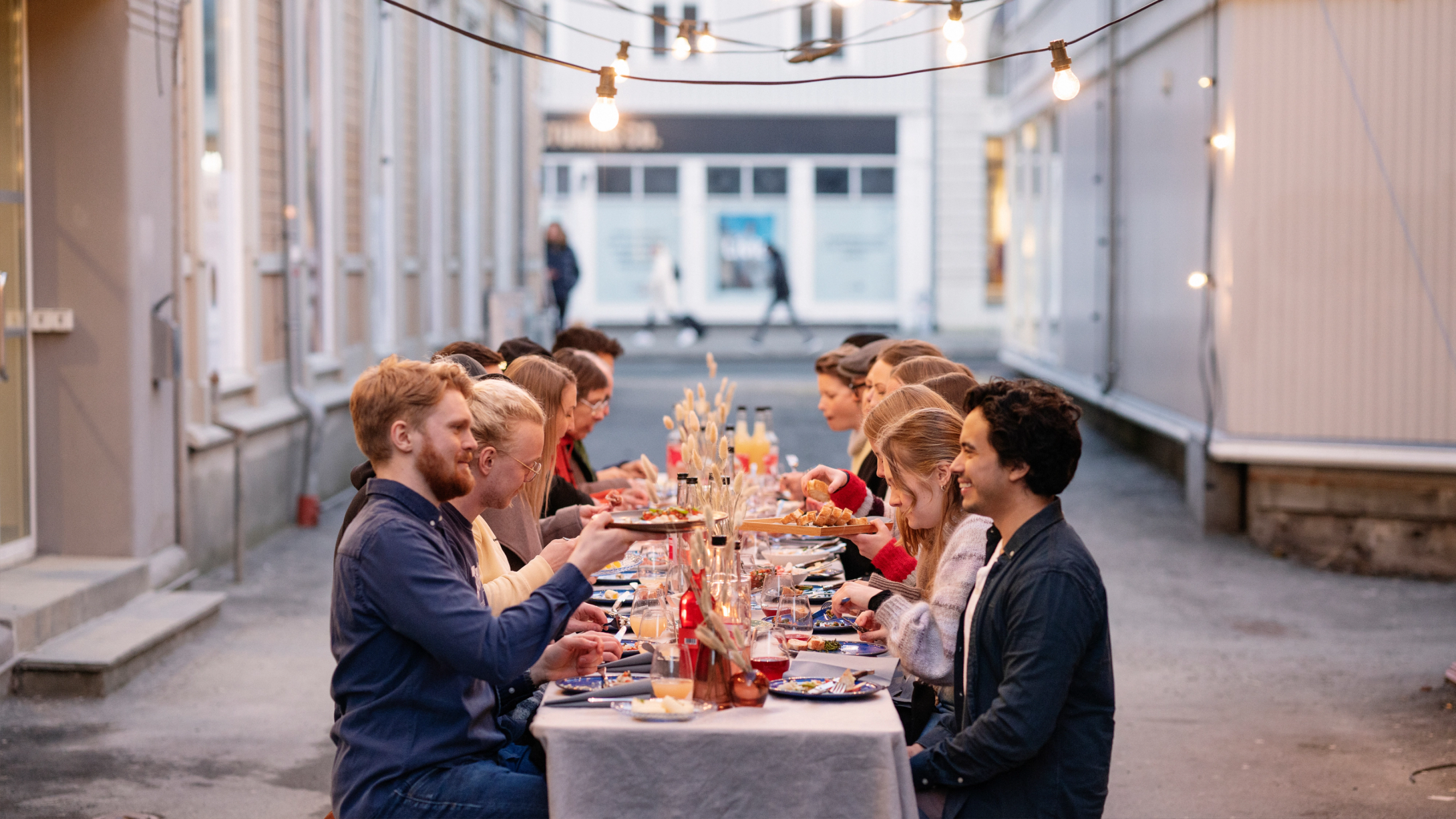 En gruppe mennesker spiser middag sammen ved et langbord i en bakgård med lyslenker over seg — av Nonspace