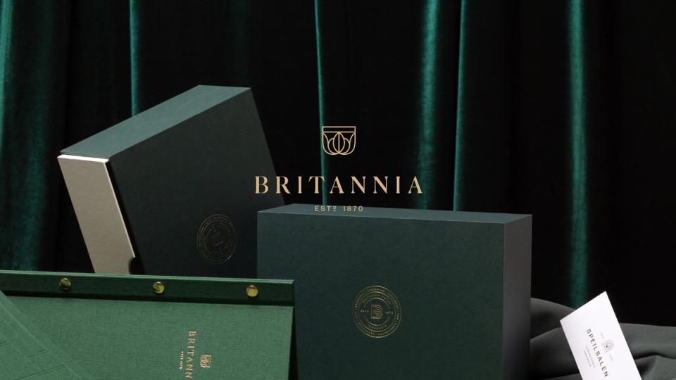 Elegante grønne gavebokser med Britannia-logo og gulldetaljer mot et mørkegrønt fløyelsbakgrunn — av Nonspace