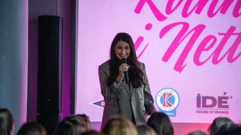 En kvinne i grå blazer holder en mikrofon og smiler på scenen foran et rosa banner med sponsorlogoer — av Nonspace