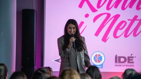 En kvinne i grå blazer holder en mikrofon og smiler på scenen foran et rosa banner med sponsorlogoer — av Nonspace