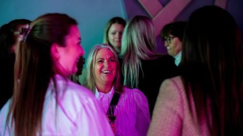 En smilende kvinne med vinglass omgitt av andre kvinner i rosa belysning på et nettverksarrangement — av Nonspace